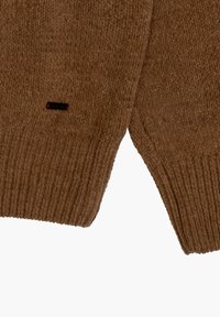 Brązowy sweter dziany o rypsowym wzorze, z kontrastującą małą czarną metką z czerwonym akcentem na dolnym szwie.