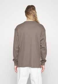 Hnědá košile s dlouhým rukávem v žebrované textuře s volným střihem a kulatým výstřihem, s rovnou spodní hranou a logem ve spodní části.