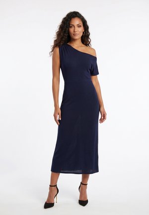 Femme aux longs cheveux bouclés portant une robe midi bleu marine à une épaule et des chaussures à talons noirs, debout devant un fond blanc.