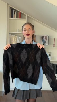 Zwart en grijs trui met ruitpatroon van gestructureerde gebreide stof, met lange mouwen en een ronde hals, vastgehouden door een persoon.