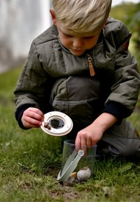 Bambino inginocchiato sull'erba, che tiene una lente di ingrandimento sopra un contenitore trasparente con conchiglie e pietre. Indossa una giacca trapuntata verde.