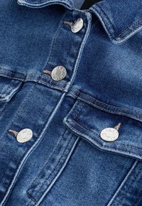 Gros plan sur le devant d'une veste en denim bleu avec des boutons métalliques étiquetés "Next Denim" et des coutures visibles sur les poches et les coutures.