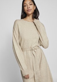 Stickad cardigan i beige med en avslappnad passform, långa ärmar, ribbade muddar och ett självsnörande midjebälte för justering. Slät textur.