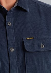 Majica tamnoplave boje s teksturom, s prednjim kopčanjem na gumbi, džepom na prsima i PM Legend logotipom. Ima okrugle gumbe i ovratnik.