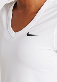 Osoba w białej sportowej koszulce Nike z dekoltem w serek, z czarnym logo swoosh po lewej stronie klatki piersiowej, ukazująca górną część torsu i linię dekoltu.