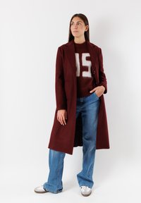 Cappotto bordeaux con un solo bottone, abbinato a un maglione rosso peloso con scritte bianche e jeans a zampa d'elefante blu chiaro. Sneakers bianche.