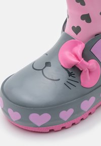 Stivale di gomma grigio da pioggia con design di un gatto sorridente, accentato da un fiocco rosa e motivi a cuore intorno alla suola, con fondo texturizzato rosa.