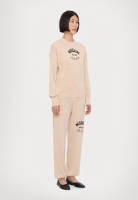 Conjunto de roupa de lazer em bege, composto por um pulôver com gola redonda e calças de corte solto. Detalhe do logótipo "Moschino Jeans" em preto e textura suave.