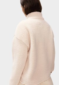 Jasnokremowa polarowa kurtka z wysokim kołnierzem, luźnym krojem i fakturowanym materiałem, z opadającymi ramionami i lekko zaokrąglonym dołem.