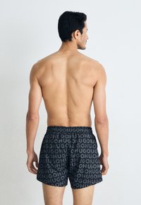 Pantaloni de baie negri cu un model repetat al logo-ului "HUGO", având o talie elastică. Partea din spate, expusă, a unui model masculin întors cu fața de la cameră.