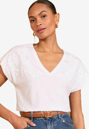White V-neck top with short sleeves, featuring floral embroidery on the shoulders. Worn with a brown belt and denim jeans.