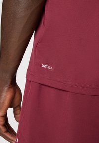 Bordeaux sporttop met een gladde textuur, voorzien van het label "DRYCELL". Close-up toont fijne naden en een getailleerd ontwerp.