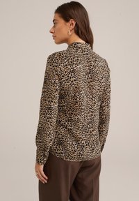 Halenka s leopardím potiskem v odstínech béžové a černé, s dlouhými rukávy, vysokým výstřihem a hladkou texturou. V kombinaci s tmavě hnědými kalhotami.
