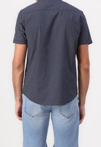 Camisa de manga corta azul marino con un patrón geométrico blanco. Tejido de algodón, dobladillo curvo y cuello abotonado. Combinada con vaqueros azul claro.