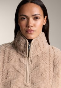 Bež pulover iz flisa z visokim ovratnikom, z zadrgo in teksturiranim vzorcem. Mehka, puhasta tkanina z gladkim detajlom zadrge.