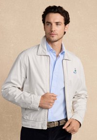 Hombre con cabello oscuro que lleva una chaqueta de cremallera color beige claro sobre una camisa de botones azul claro, ajustándose la chaqueta cerca del cuello.