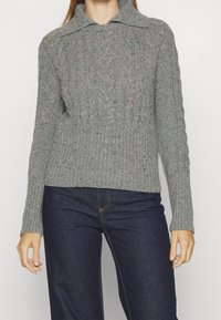 Grå stickad tröja med krage, med en ribbad fåll och en texturerad flätad mönster. Flerfärgade inslag genom hela tyget.