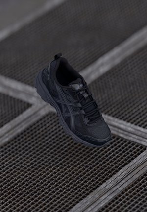 Chaussure de sport noire avec une tige texturée, design à lacets, col rembourré et semelle cushioned ; présente le logo Asics sur la languette.