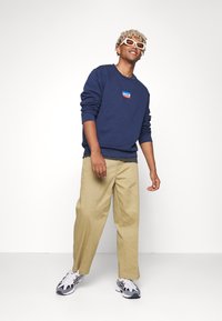 Marinblå sweatshirt med en logotyp på framsidan, tillsammans med beiga byxor. Vita sneakers med grå detaljer. Modellen står med ett litet leende.