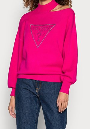 Femme portant un pull rose vif avec le logo "GUESS" en strass et un jean bleu, visible du cou jusqu'aux hanches.