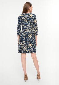 Dunkelblaues Kleid mit Dreiviertelärmeln, das mit einem beigen Blumenmuster verziert ist. Glattes Material, knielanger Schnitt, kombiniert mit nudefarbenen hohen Schuhen.
