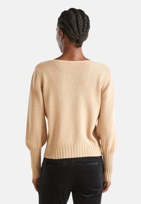 Person mit geflochtenen Haaren, die einen beigen Strickpullover und schwarze Hosen trägt, steht mit dem Rücken zu einem schlichten, hellen Hintergrund.