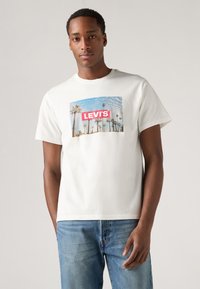 Tricou alb din bumbac cu un grafic rectangular care prezintă un design cu palmier și textul "LEVI'S" în roșu. Croială lejeră, mâneci scurte.