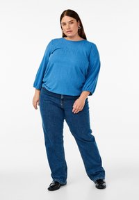 Blau strukturierter langärmliger Oberteil, kombiniert mit dunkelblauen Jeans. Das Oberteil hat einen runden Ausschnitt und leicht gepuffte Ärmel. Schwarze Schuhe vervollständigen das Outfit.