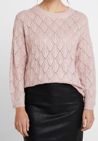 Maglione lavorato a maglia rosa chiaro con motivo a rombi, scollo rotondo e maniche lunghe, abbinato a una gonna nera in pelle a vita alta.
