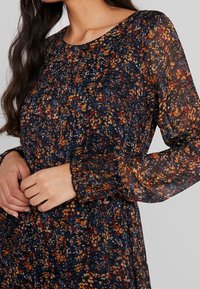 Šaty s dlouhým rukávem, které mají průhledný, vícebarevný květinový vzor na tmavém pozadí. Rukávy jsou na koncích nařasené pro strukturovaný vzhled.