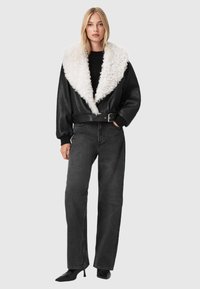 Sort læderjakke med en hvid falsk pelskrave, bælte i taljen og lange ærmer, parret med højtaljede sorte jeans og spidse hæle.