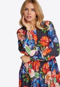 Blumenkleid mit lebhaften roten, pinken und blauen Blumen auf einem dunklen Hintergrund. Geraffte Smocking-Details an den Bündchen und in der Taille. Lange Ärmel.