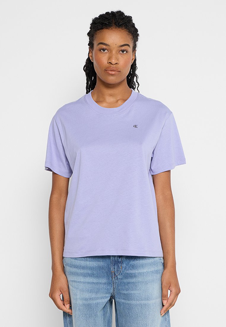 Kobieta stoi przodem, ubrana w jasnofioletową koszulkę Calvin Klein i jasnoniebieskie dżinsy, na tle gładkiego, białego tła.