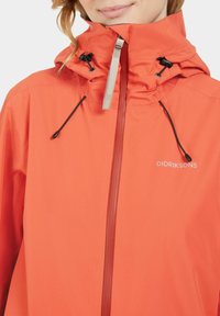 Heldere oranje regenjas met een hoge kraag, verstelbare capuchon, zwarte koordjes en een zilveren rits. Voorzien van een logo op de borst.