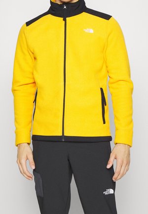 Homme portant une veste polaire jaune avec des accents noirs et des poches zippées, associée à un pantalon noir avec une poche latérale.