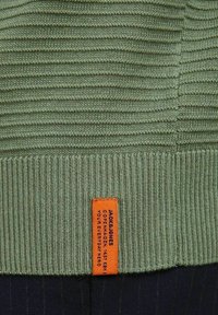 Groene ribgebreide stof met een oranje rechthoekig kledinglabel genaaid aan de onderrand, met zwarte bedrukte merktekst.