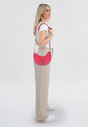 Borsa a tracolla in pelle rosa con design a cuore trapuntato, tracolla in catena argentata e hardware nero, indossata sul fianco di un modello in pantaloni beige.