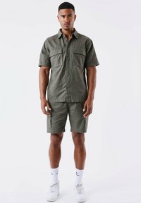 Camisa de manga corta verde oliva con dos bolsillos frontales, combinada con pantalones cortos cargo a juego. La tela parece ligera con un acabado texturizado.