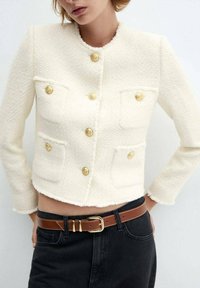 Veste blanche en tweed raccourcie avec quatre poches devant, boutons dorés et ourlet effiloché ; portée avec un pantalon noir et une ceinture marron.