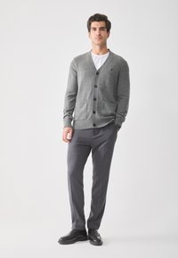 Cardigan de lana gris con botones frontales y bolsillos, combinado con pantalones gris oscuro y zapatos negros, llevado por una persona de pie.
