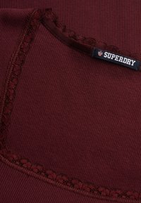 Bordeauxrødt ribbet stof med blonderkant omkring en firkantet halsudskæring og et sort mærke med teksten "SUPERDRY" syet i øverste kant.