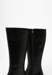 Bottes montantes noires en cuir lisse, dotées d'une fermeture éclair latérale et de coutures minimales, présentant des éléments de design modernes.