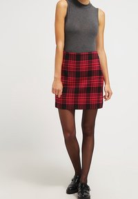 Zwart-rode geruite minirok met een aansluitende taille, gecombineerd met een grijs mouwloos topje en ondoorzichtige zwarte panty's. Platte zwarte schoenen.