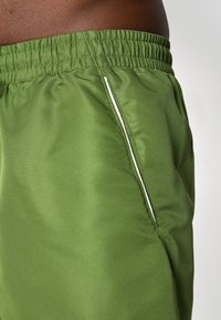 Calções de desporto verdes feitos de um tecido suave e leve; apresentam uma cintura elástica e um bolso com fecho éclair branco. Padrão texturizado fino.