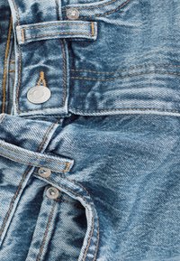 Denimo audinys, rodantis artimą mėlynųjų džinsų vaizdą su matomomis siūlėmis, sagčių užsegimais ir tekstūruota paviršiumi. Akiniai iš aukso siūlų.