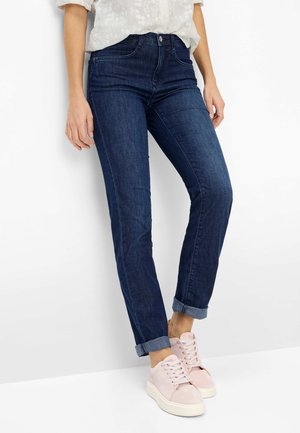 Frau trägt dunkle, umgeschlagene Jeans und hellrosa Sneaker, steht vor weißem Wand- und Fußbodenhintergrund.