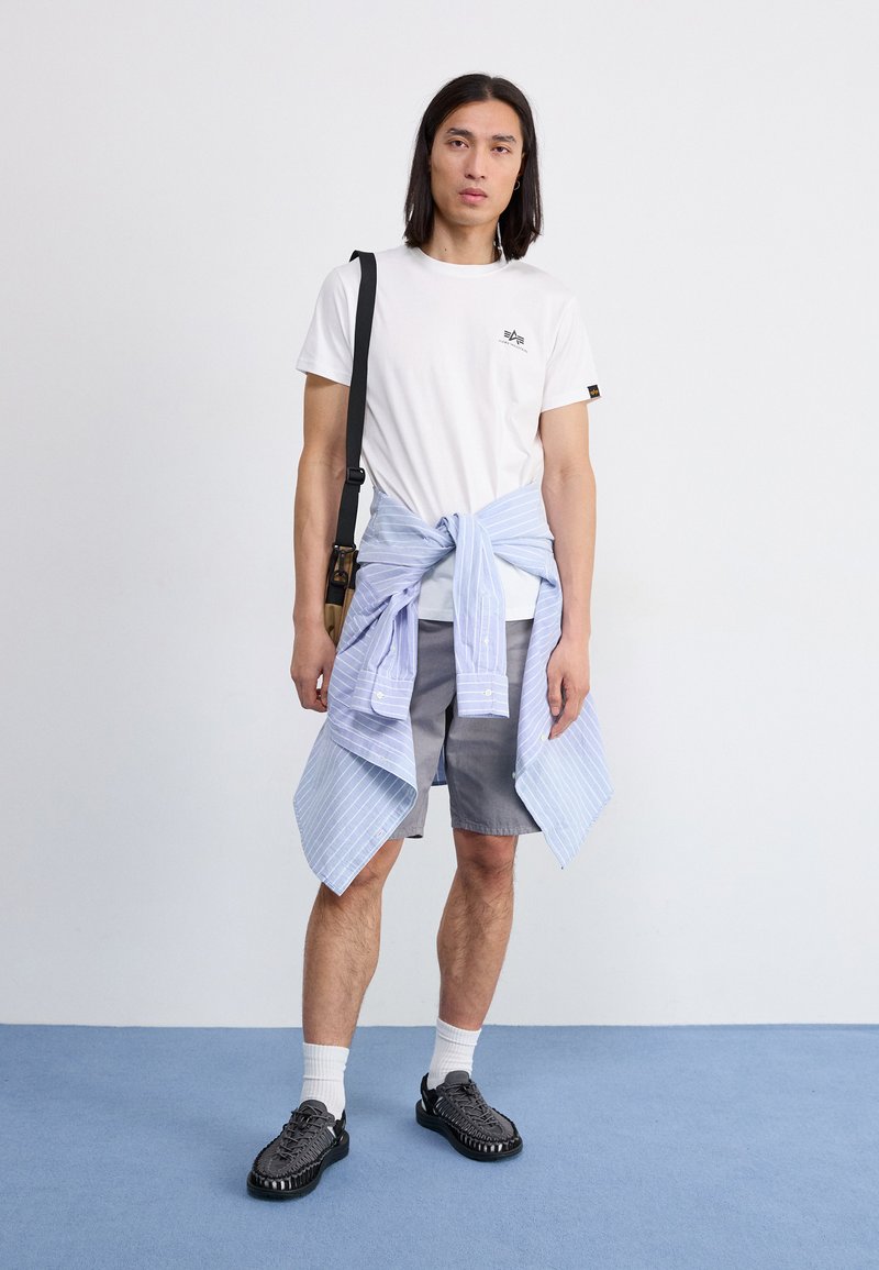 Weißes T-Shirt mit Logo, graue Shorts und ein hellblau gestreiftes Hemd um die Taille gebunden. Schwarze Schuhe und weiße Socken. Einfaches Design.