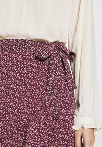 Skirt s květinovým vzorem v burgundské barvě, se zavázáním v pase, spárovaná s krémovou blůzou s volánovými manžetami. Měkká látka a jemné detaily vzoru.