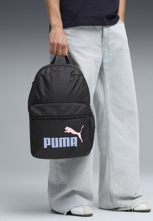 Personne tenant un sac à dos noir Puma par sa poignée supérieure, portant un jean large bleu clair et des baskets blanches sur fond gris.