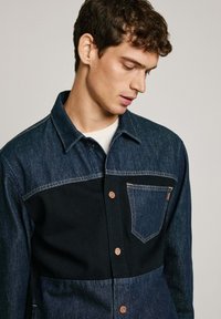 Camicia di denim con una parte superiore blu scuro e una parte inferiore nera, dotata di taschino frontale, cuciture in evidenza e bottoni di rame.